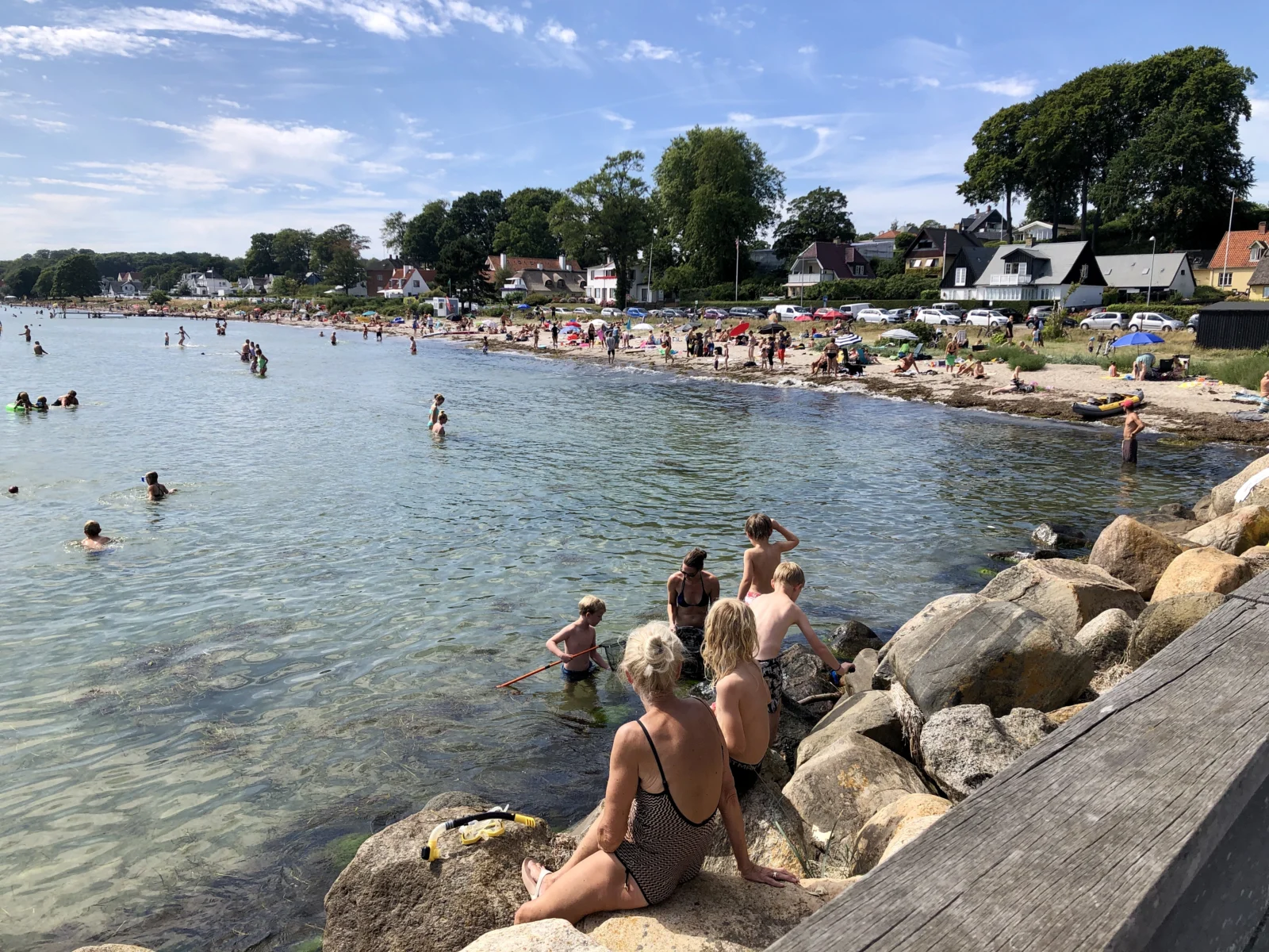 Badegæster ved Snekkersten Strand med grønne træer og huse langs Strandvejen